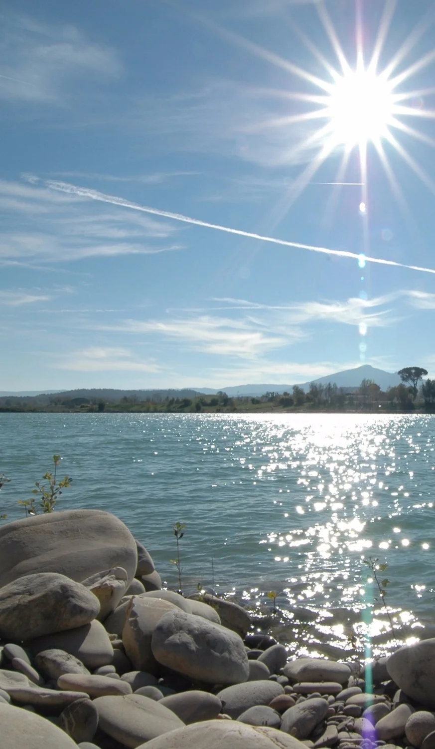 Vue ensoleillée d'un lac aux eaux scintillantes, rive de galets au premier plan, collines boisées à l'horizon sous un ciel bleu éclatant